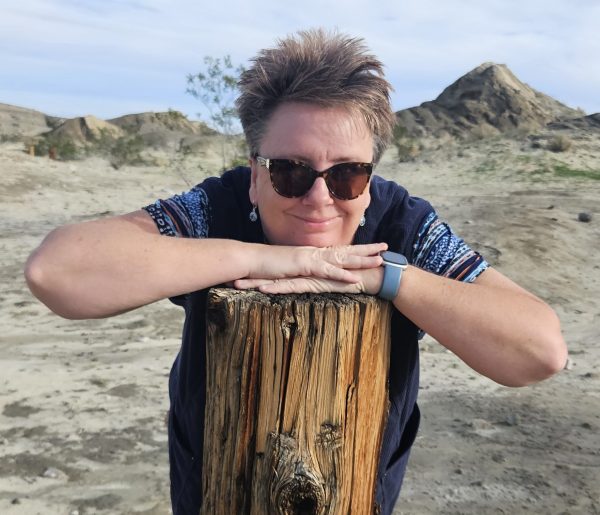 A smiling woman in sunglasses leans her chin on her hands atop a weathered wooden post, with a vast, sandy desert landscape in the background.