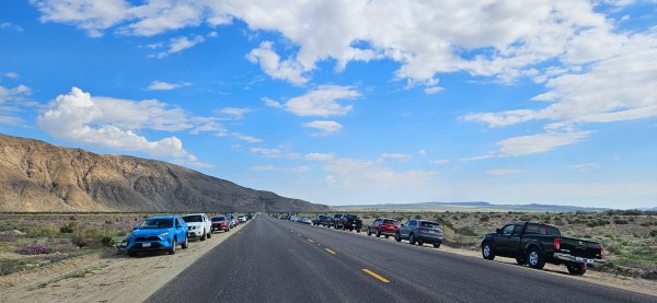 Henderson Canyon road with parked cars lining one side, under a blue sky with scattered clouds and mountains in the background.